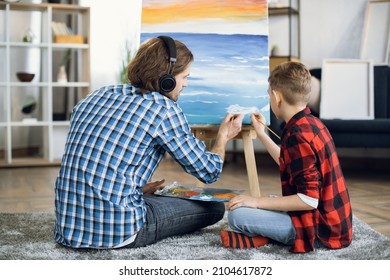 Back View Of Caucasian Man And Boy Wearing Checkered Shirts Using Brushes And Color Palette For Painting On Canvas At Home. Father And Son Sitting On Floor And Enjoying Creative Process.