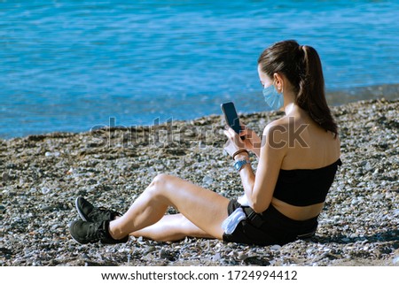 back view of caucasian millennial female seated on sandy beach next to the sea in sport clothes, consulting cellphone and wearing face mask. Concept for vacation, sport