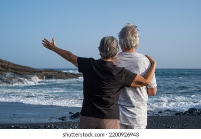 Back View Of Caucasian Senior Couple Standing On The Beach At Sunset Looking At Horizon Over Water - Relaxed Pensioners Enjoying Free Time, Sea Vacation Or Retirement