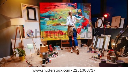 Back view of Caucasian young talented female painter working in cozy workshop in evening emotionally drawing on big canvas colorful abstract painting. Artwork, contemporary oil painting, work concept