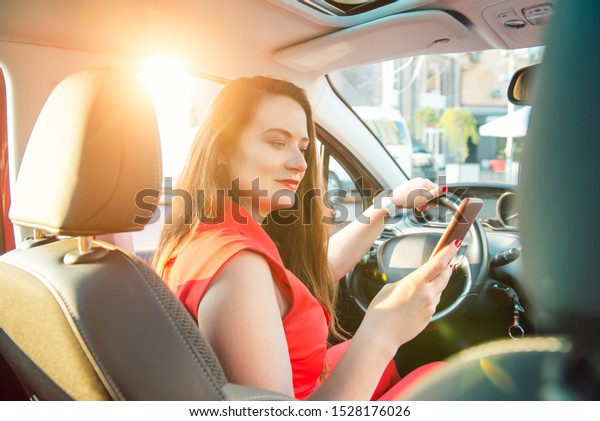 Back view\
caucasian young woman driver using touch screen smartphone and hand\
holding steering wheel in a car during driving. Sunset back light.\
Selective focus, copy space. Inside\
view