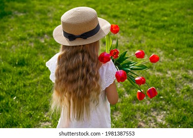 Back view from the back of a child girl with long blond golden wavy hair in a hat, against a background of green grass lawn, holding a bouquet of red tulips.