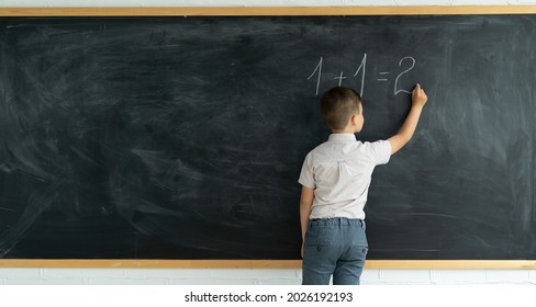 Back view of a child writes with chalk on a blackboard solution of an example in mathematics. Elementary school math lesson. Schoolboy boy thinks near the chalkboard. Student at the lesson. - Shutterstock ID 2026192193