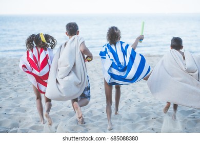 Back View Of Children With Towels As Superhero Robes And Water Toys In Hands Running On Beach