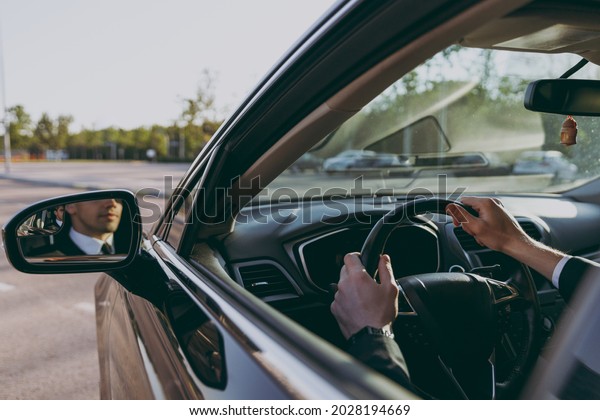 Back view close up cropped up photo young driver\
reflected in mirror man wearing black suit driving car taxi hold\
steering wheel look camera Vehicle transport traffic lifestyle\
business trip concept.