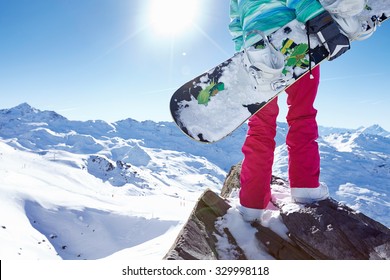 Back view close up of female snowboarder wearing helmet, blue jacket, grey gloves and pink pants standing with snowboard in one hand and enjoying alpine mountain landscape - winter sports concept