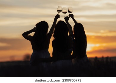 Back view of company of young female friends with hands up having fun, raising glasses with wine and enjoy beautiful sunset at summer picnic in lavender field.	
 - Shutterstock ID 2309900993