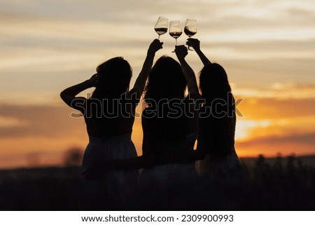 Back view of company of young female friends with hands up having fun, raising glasses with wine and enjoy beautiful sunset at summer picnic in lavender field.	
