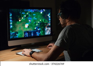 Back view of concentrated young gamer in headphones and glasses using computer for playing game at home
