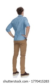 back view of concerned casual guy holding hands on hips and thinking, standing isolated on white background, full body