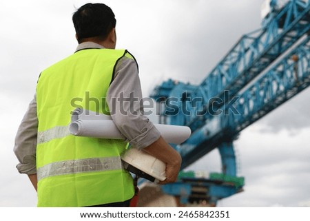 back view of construction engineer in green vest safety wear and white helmet holding blueprint. isolated on a huge crane bridge building background