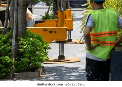 Back view of a construction worker in a bright green safety vest with orange stripes, standing near yellow hydraulic outriggers of a heavy vehicle deployed on a paved sidewalk.