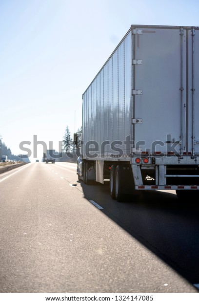 Back view of convoy of professional logistic freight\
transportation commercial big rigs semi trucks running with\
different semi trailers on the straight divided highway road\
towards the sun