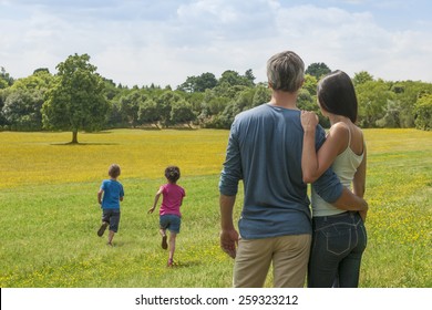 Back View Of A Couple Holding Each Other And Contemplating A Landscape While Their Two Children Running In The Grass