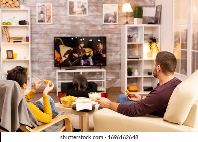 Back View Of Couple In Living Room Watching A Movie On The TV While Eating Takeaway Food