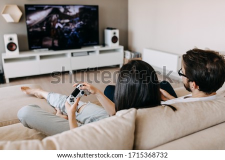 Back view of a couple of man and woman playing video game on a console, sitting on couch in living room.
