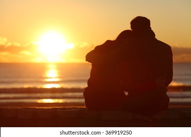 Back view of a couple silhouette watching sun at sunset on the beach in winter with a warmth light