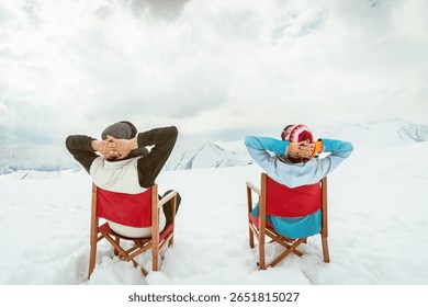 Back view couple sitting in deck chairs on snow hands behind heads enjoying mountain view winter leisure