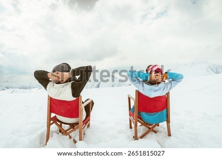 Back view couple sitting in deck chairs on snow hands behind heads enjoying mountain view winter leisure