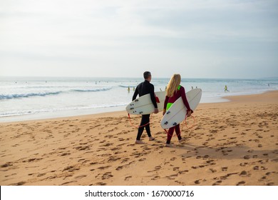 Back View Of Couple With Surfboards Walking On Beach. Rear View Of Man And Woman In Wetsuits Holding Surfboards And Walking On Sea Coast At Summertime. Surfing Concept