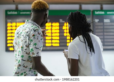 Back view of couple in a train station wearing protective face masks, looking at their smartphone and the travel board with train schedule information.