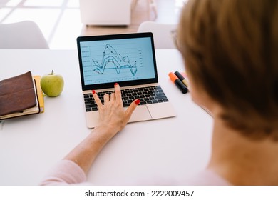 Back View Of Crop Anonymous Woman Pointing At Screen Of Laptop While Sitting At Table With Notepad Markers And Apple Fruit