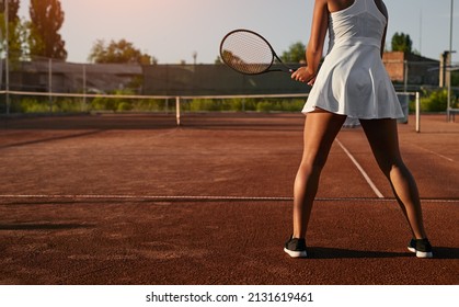 Back view of crop sportswoman in white dress grasping racket tight while preparing to play tennis on court at sunset