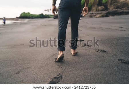 Back view of crop unrecognizable barefoot person walking along wet sandy beach against blurred background while enjoying vacation alone on seashore