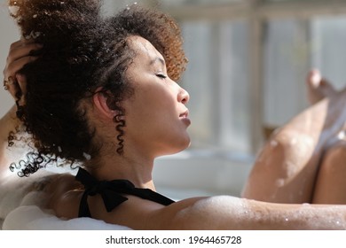back view curly black girl laying, relaxing in the bath indoors. young woman relaxing in warm bathtub with foam and bubbles. Tired female enjoying rest pamper herself.
