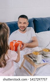 Back View Of Cute Child Presenting Fathers Day Gift Box To Smiling Father Having Breakfast In Bed