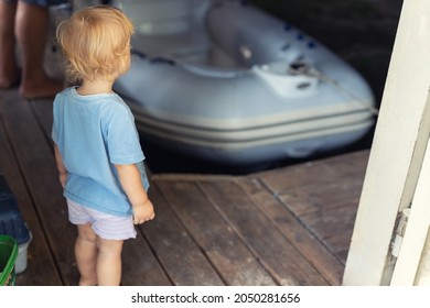 Back View Of Cute Little Caucasian Toddler Boy Stand Alone In Ship Garage Wooden Floor Dock And Looking On Inflatable Boat Dreaming Of Sailing By Lake Or Pond River. Summer Outdoor Activity