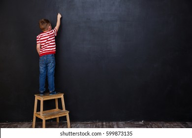 Back view of cute little kid boy standing on ladder and writing with chalk on the backboard in school class - Powered by Shutterstock