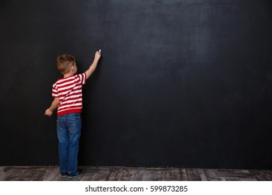 Back View Of Cute Little Kid Boy Writing With Chalk On The Backboard In School Class