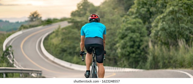 back view of a cyclist on top of a mountains winding road, riding a black bicycle down a hill, wearing bike helmet and blue cycling jersey, with grey clouds sunset sky and forest ,banner