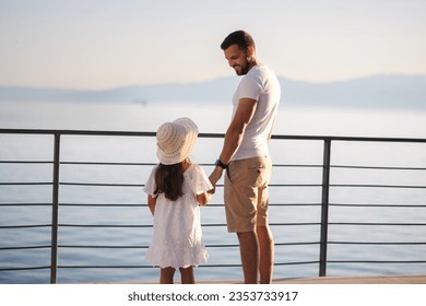 Back view of daughter with her father stand on heigh balcony in front of sea sunset. Dad with his little girl looking on mountains