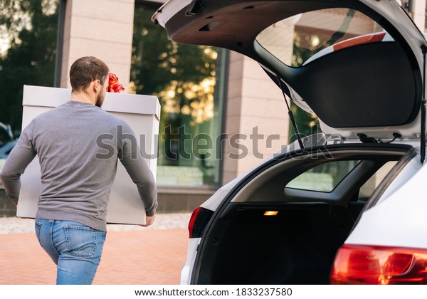 Back view of delivery man takes out large\
festive white box with beautiful red bow from trunk of car for\
delivery to client, outdoors. Courier preparing to take parcel out\
and deliver it to\
customer.