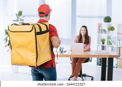 Back View Of Delivery Man With Thermo Backpack And Smiling Businesswoman At Table In Office