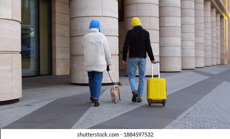 Back View Of Diverse Couple In Knitted Hat And Warm Clothes Walking With Luggage And Pet Dog Outside Airport. Multiethnic Man And Woman Traveling Together With Pug Dog