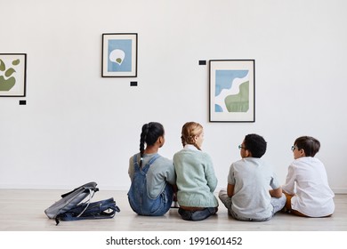 Back view at diverse group of children sitting on floor in modern art gallery and discussing paintings, copy space