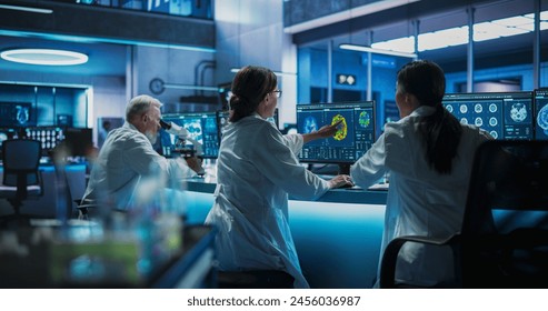 Back View Of Diverse Scientists Working In Medical Research Center. Male Doctor Using Microscope And His Female Colleagues Looking At MRI Brain Scans On Desktop Computers. Biotechnology Lab Concept.