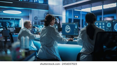 Back View Of Diverse Scientists Working In Medical Research Center. Male Doctor Using Microscope And His Female Colleagues Looking At CT Brain Scans On Desktop Computers. Biotechnology Lab Concept.