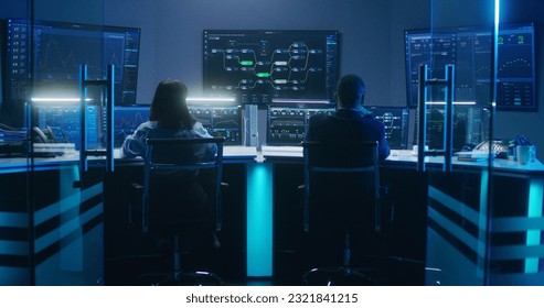 Back view of diverse IT technical specialists working on computers with database server in modern monitoring office at night. Big digital screens with real-time analysis charts and blockchain network.