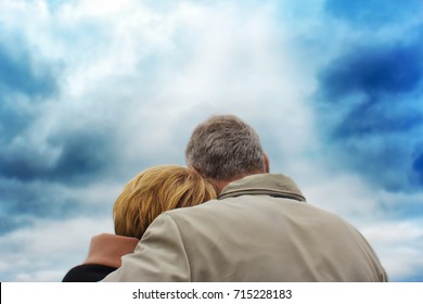 Back View To An Elderly Couple. The Woman Put Her Head On The Man's Shoulder. Against A Background Of A Deep Blue Sky.