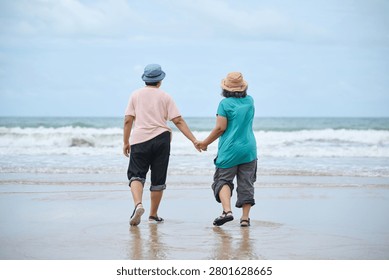 Back view elderly people walking on the beach hand-in-hand, facing the ocean with gentle waves lapping at the shore