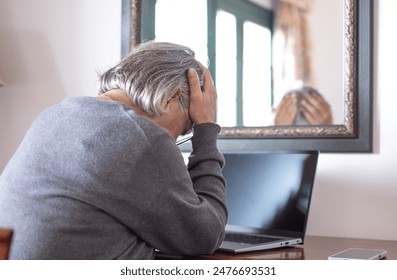 Back view of elderly woman using laptop at home while touching her head for headache. Excessive work or study generates severe migraine