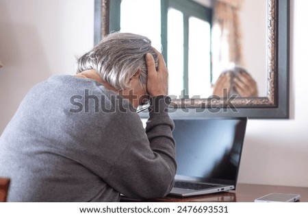 Back view of elderly woman using laptop at home while touching her head for headache. Excessive work or study generates severe migraine