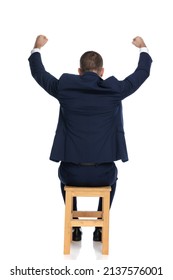 back view of elegant businessman in suit with hands above head sitting on wooden chair and celebrating victory on white background in studio