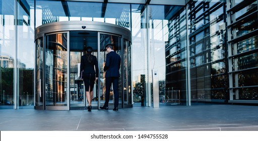 Back View Of Elegant Man And Woman Walking Into Round Revolving Doors While Entering Contemporary Glass Skyscraper Building Of Corporation Office