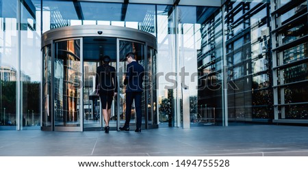 Back view of elegant man and woman walking into round revolving doors while entering contemporary glass skyscraper building of corporation office