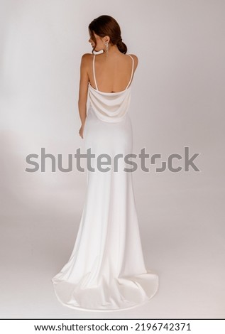Back view of elegant woman in a wedding long dress with open back posing on white background. Fashionable dress with bare shoulders. Wedding details. Slim female model. Jewelry.